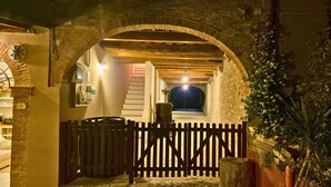 Une entrée en arc de cercle, faite de pierres, mène à un bâtiment entouré d’une clôture en bois. On y aperçoit un tonneau et un escalier qui donne sur un espace éclairé situé plus loin.