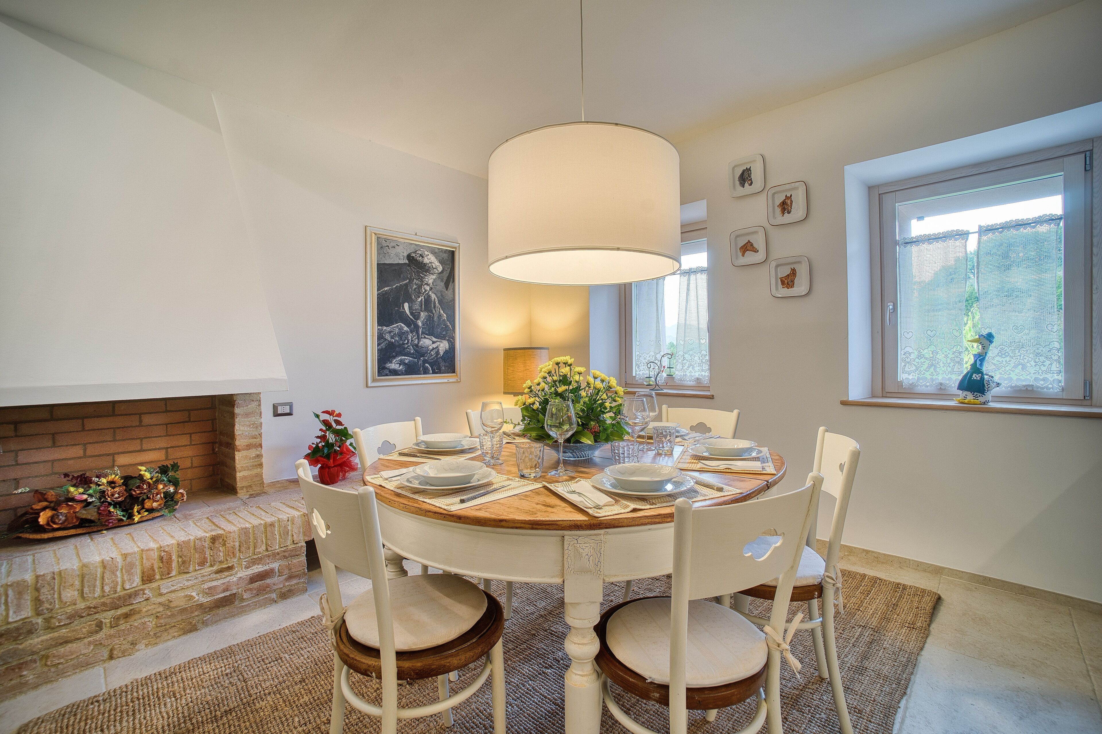 Un coin repas avec une table ronde dressée pour quatre personnes, entourée de chaises blanches. Un grand lustre est suspendu au-dessus de la table, et une cheminée avec un manteau en briques est visible sur la gauche.