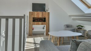 Ein moderner Loft-Wohnraum mit hohen Decken, Dachfenstern, einem eingebauten Kamin, einem Flachbildfernseher und einer gemütlichen Sitzecke mit grauem Sofa und rundem Tisch.