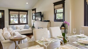 A dining area with a wooden table set for four, surrounded by white chairs. A television is mounted on the wall above the table.