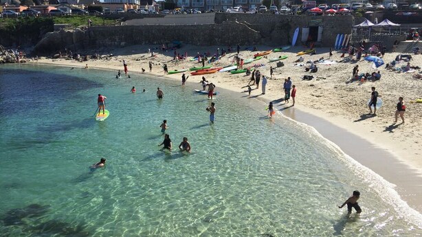 A beach with clear water, people swimming and paddleboarding, and boats on the shore.