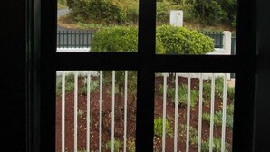 A view through a window with a metal railing, overlooking a road, greenery, and a distant shoreline under a cloudy sky.