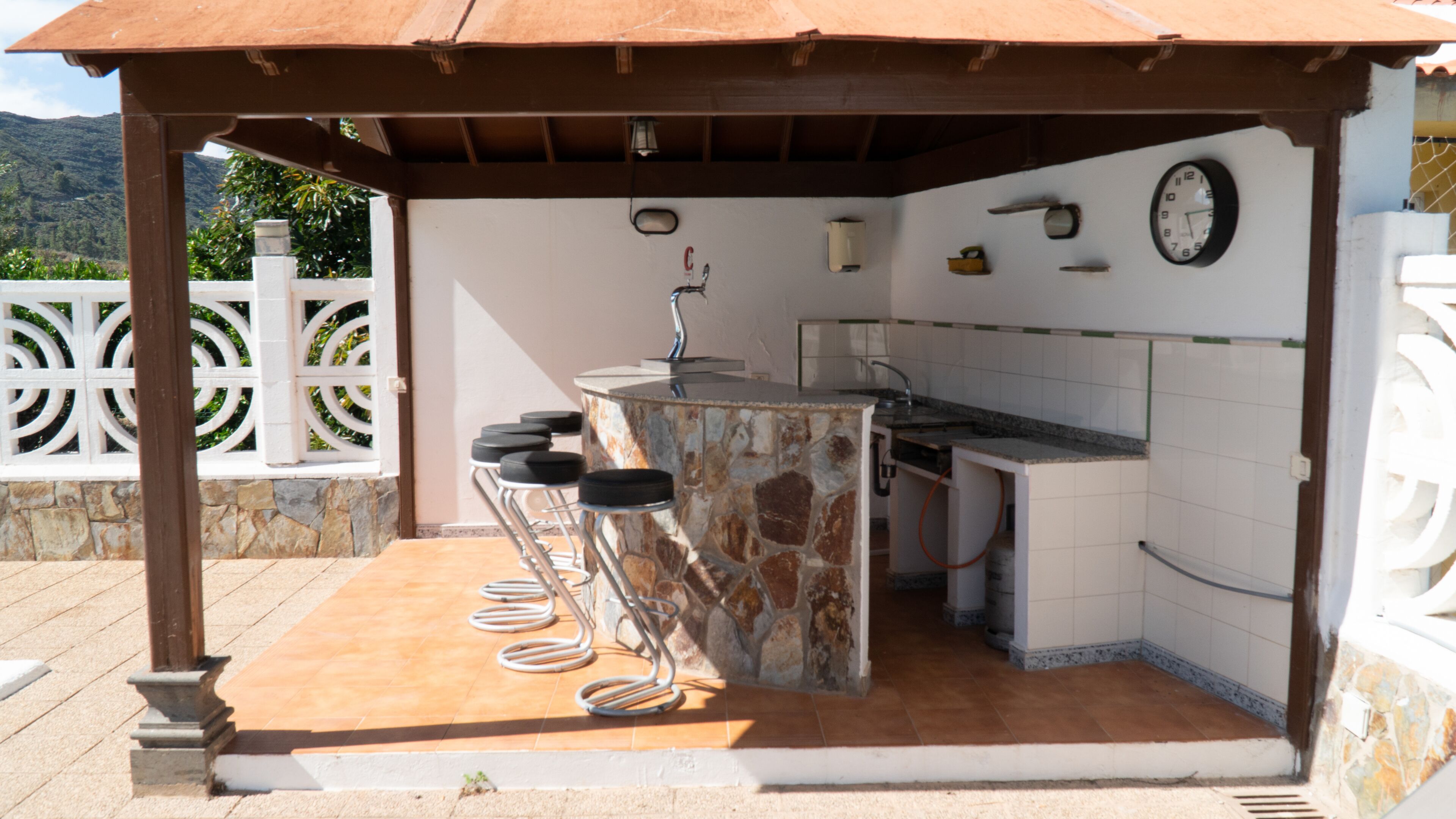 An outdoor kitchen area with a stone counter, bar stools, a sink, and a clock on the wall.