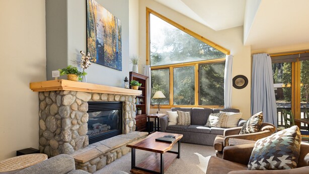 A cozy living room with a stone fireplace, comfortable seating, and large windows offering a view of the outdoors.