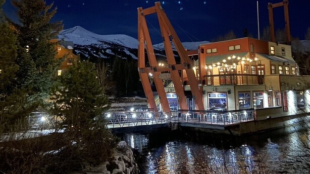 A riverfront building with a snow-capped mountain in the background, a wooden bridge, and a lit-up facade.