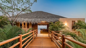 A wooden bridge leading to a thatched-roof structure with a view of the sea. - Luxe Beach Villa in Exclusive Oceanfront Community (Huatulco)