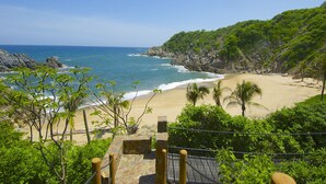 A coastal landscape with a sandy beach, rocky cliffs, and lush greenery. - Luxe Beach Villa in Exclusive Oceanfront Community (Huatulco)