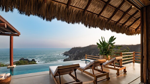 A wooden infinity pool with lounge chairs and a potted plant, overlooking a rocky coastline. - Luxe Beach Villa in Exclusive Oceanfront Community (Huatulco)