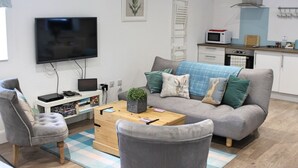 A modern living room with a grey sofa, a coffee table, and a TV. There is a kitchen area in the background with a microwave and a kettle. - A cute and cosy cottage, close to the train station. (dunblane)