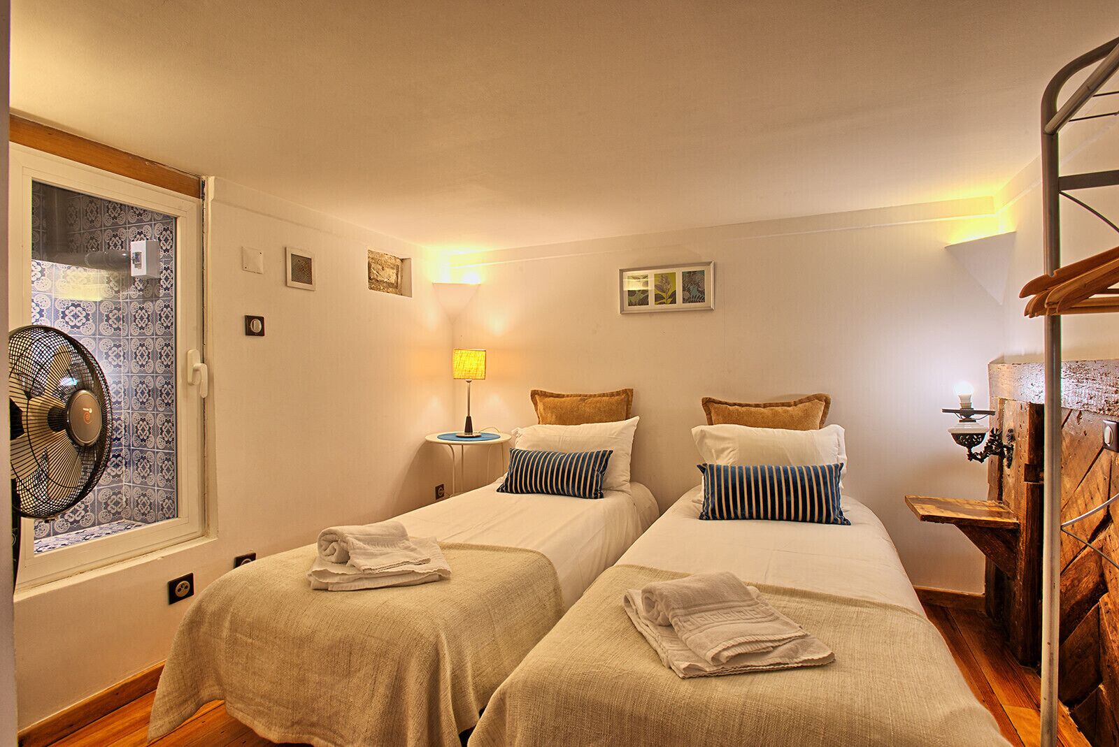 Camera d'albergo con due letti singoli, una ventola da terra, una lampada da tavolo e una scala in legno.