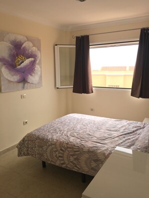 Une chambre à coucher avec un lit, une fenêtre avec des rideaux, un miroir fixé au mur et un grand tableau représentant des motifs floraux.