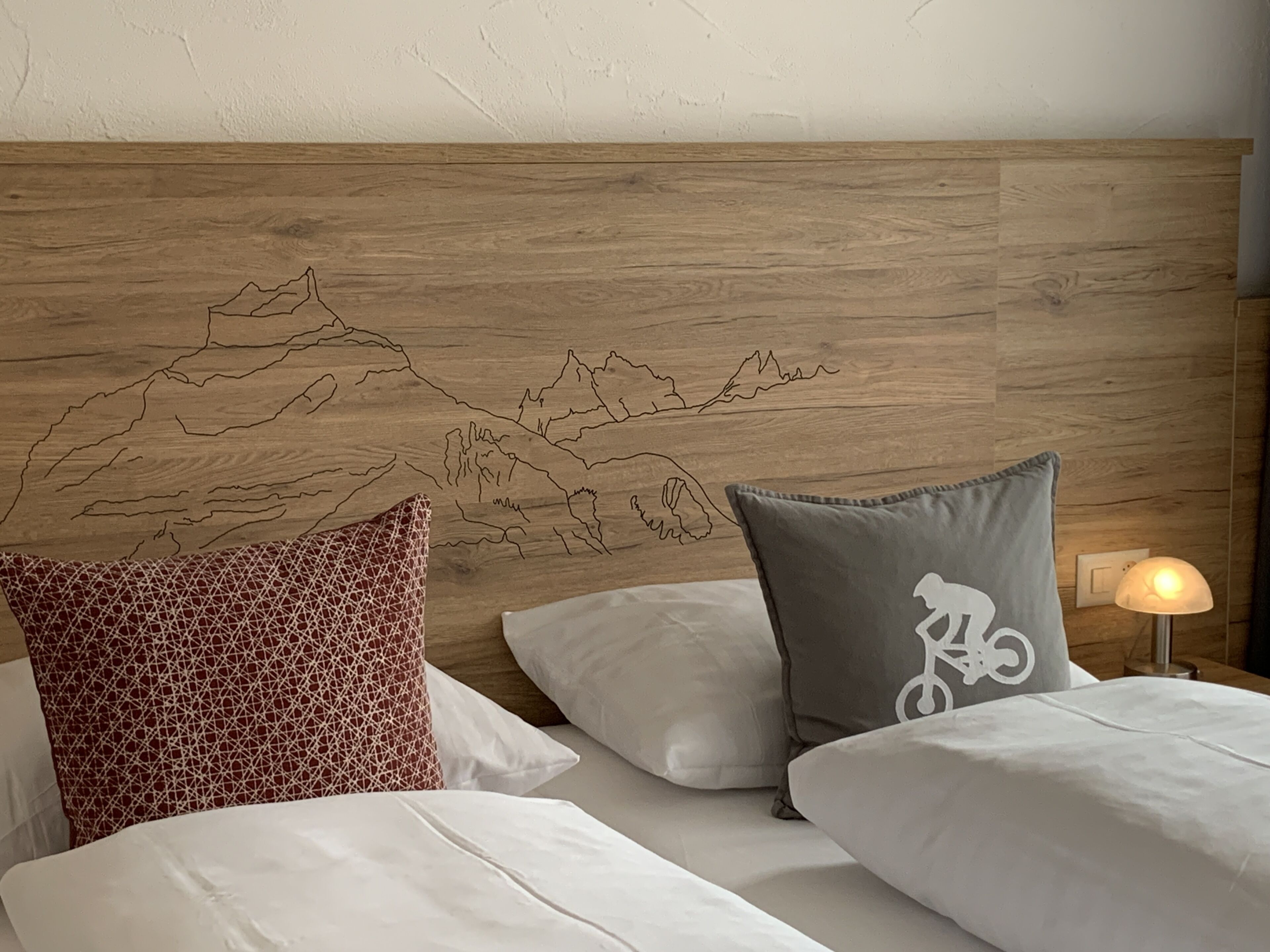 Une chambre d’hôtel avec une tête de lit en bois ornée d’un motif représentant un paysage de montagne, du linge de lit blanc et une lampe sur la table de chevet.
