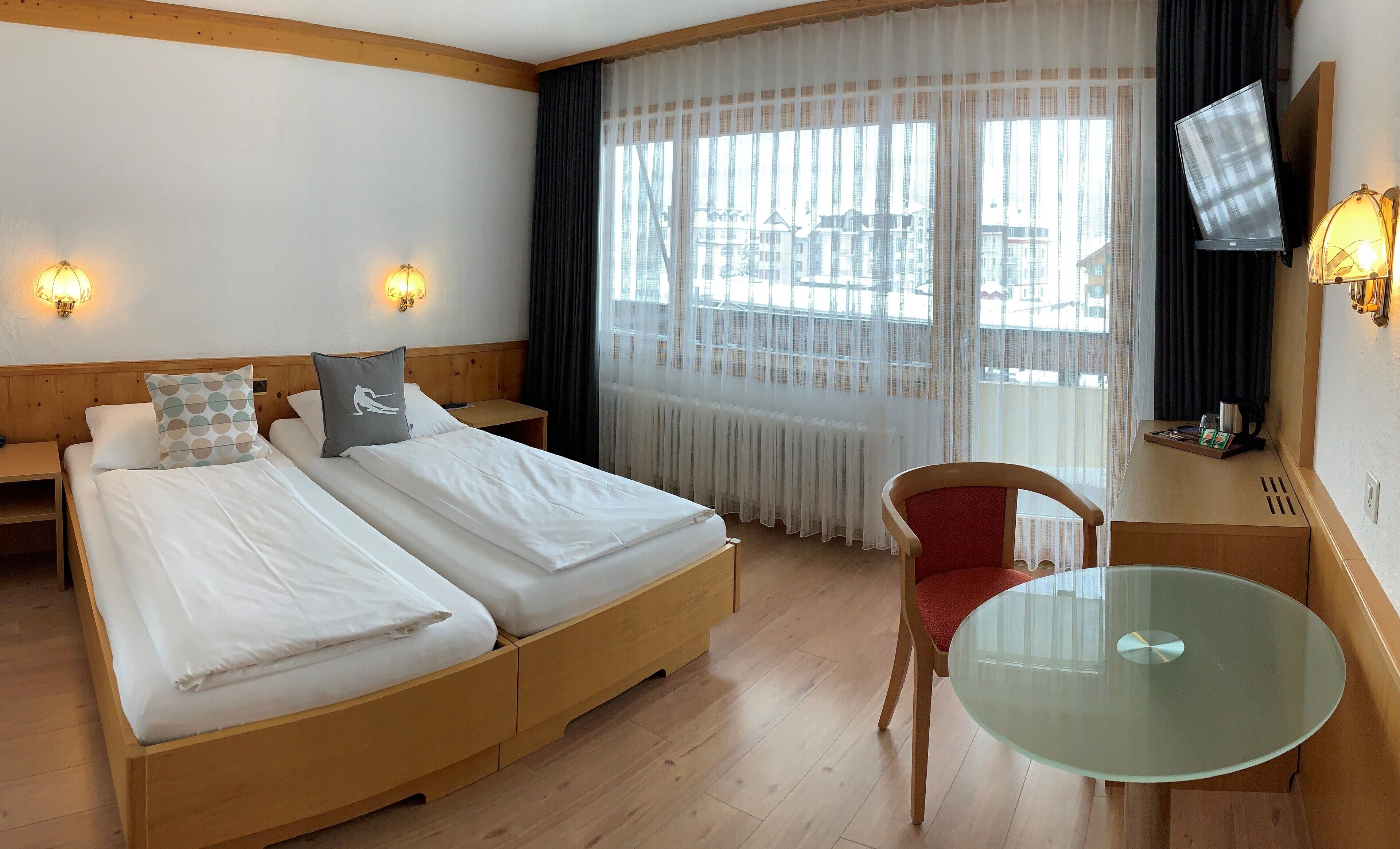 Habitación de hotel con dos camas, una mesita, una silla, televisión y ventana con cortinas traslúcidas.