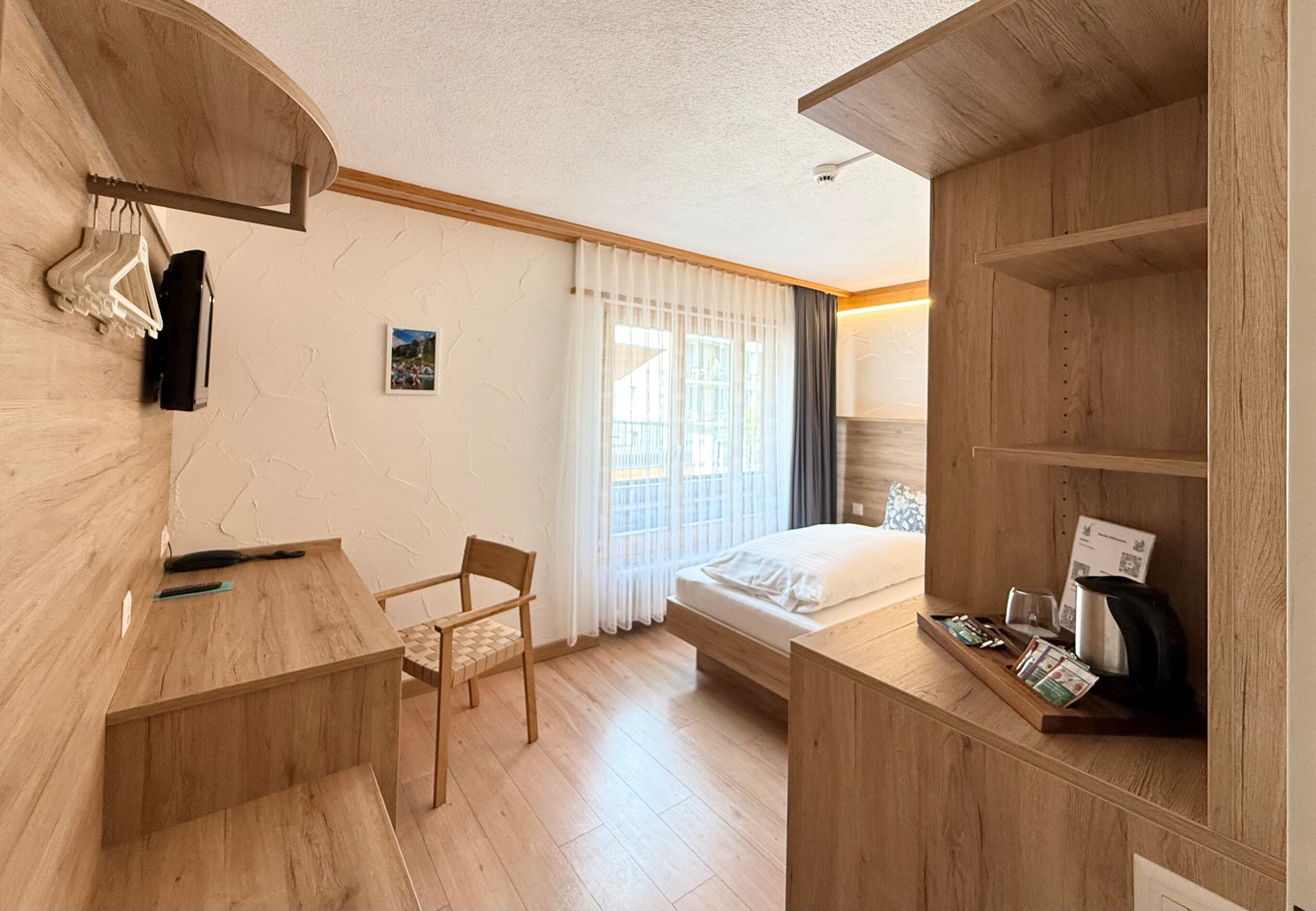 A hotel room with a wooden desk, a chair, a bed, and a shelf with a kettle and a tray of items.