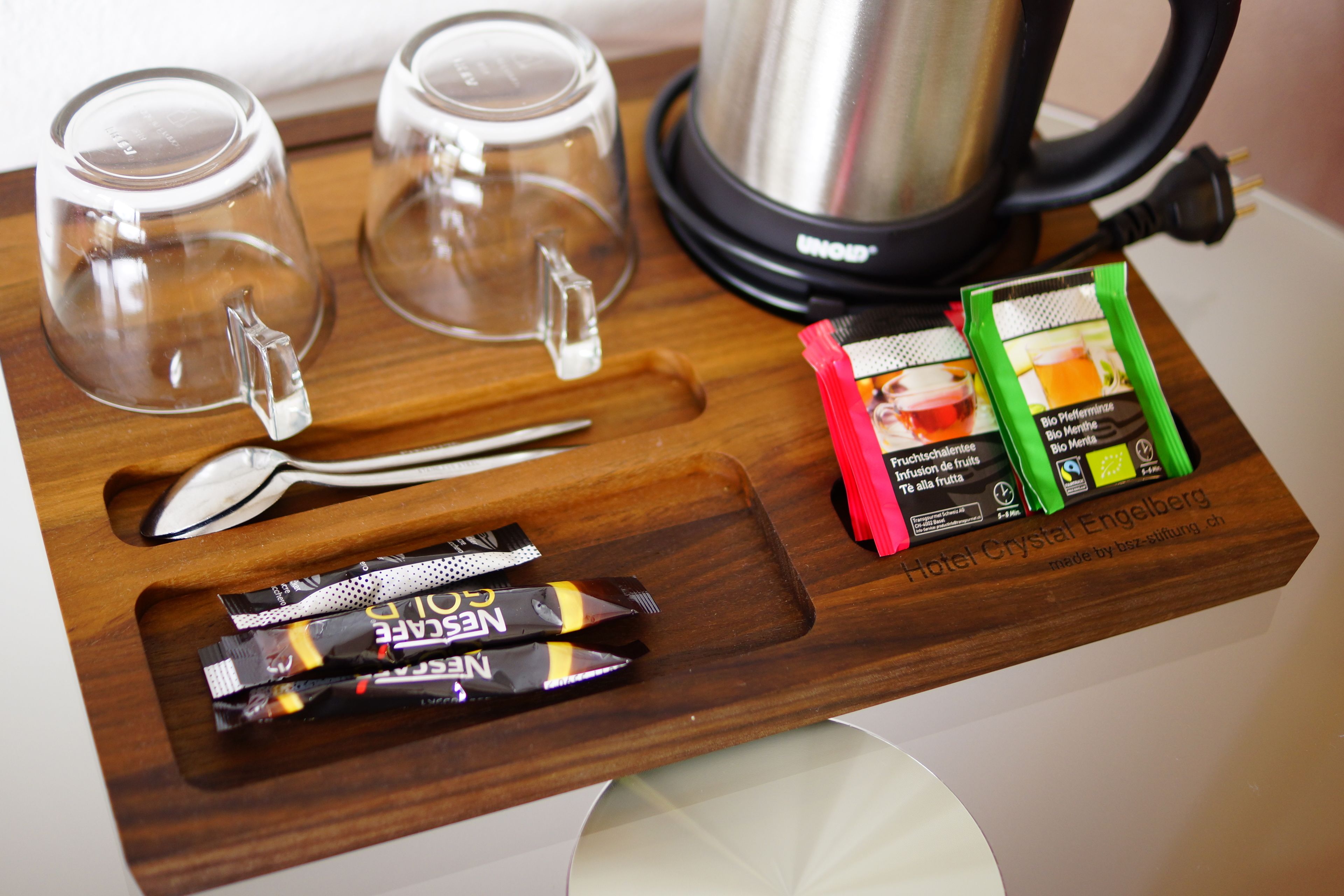Un plateau en bois avec deux verres transparents, une cuillère, deux sachets de chocolat Nestlé et un sachet de thé vert et rouge.