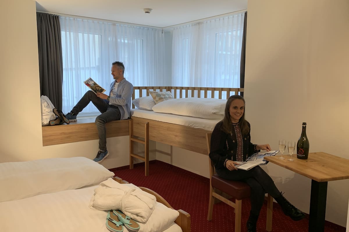 Une chambre d’hôtel avec deux lits, une table en bois sur laquelle se trouve une bouteille, et une fenêtre avec des rideaux.