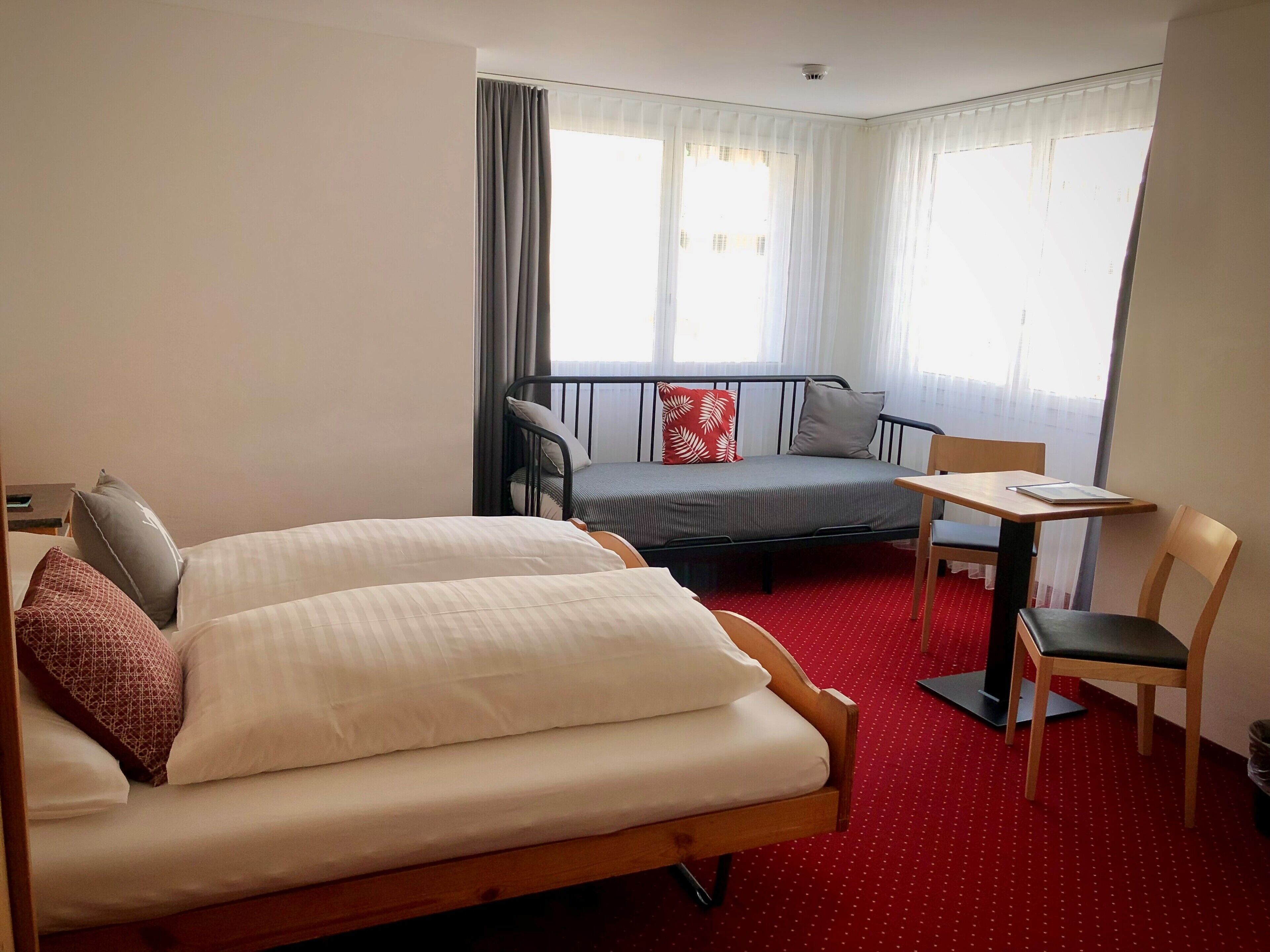 A hotel room with two beds, a small table, a chair, and a balcony with curtains.