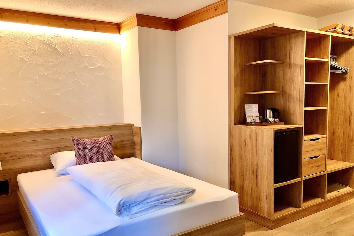 Un lit bien fait, avec une tête de lit en bois, une armoire en bois dotée d’étagères et un luminaire fixé au mur.
