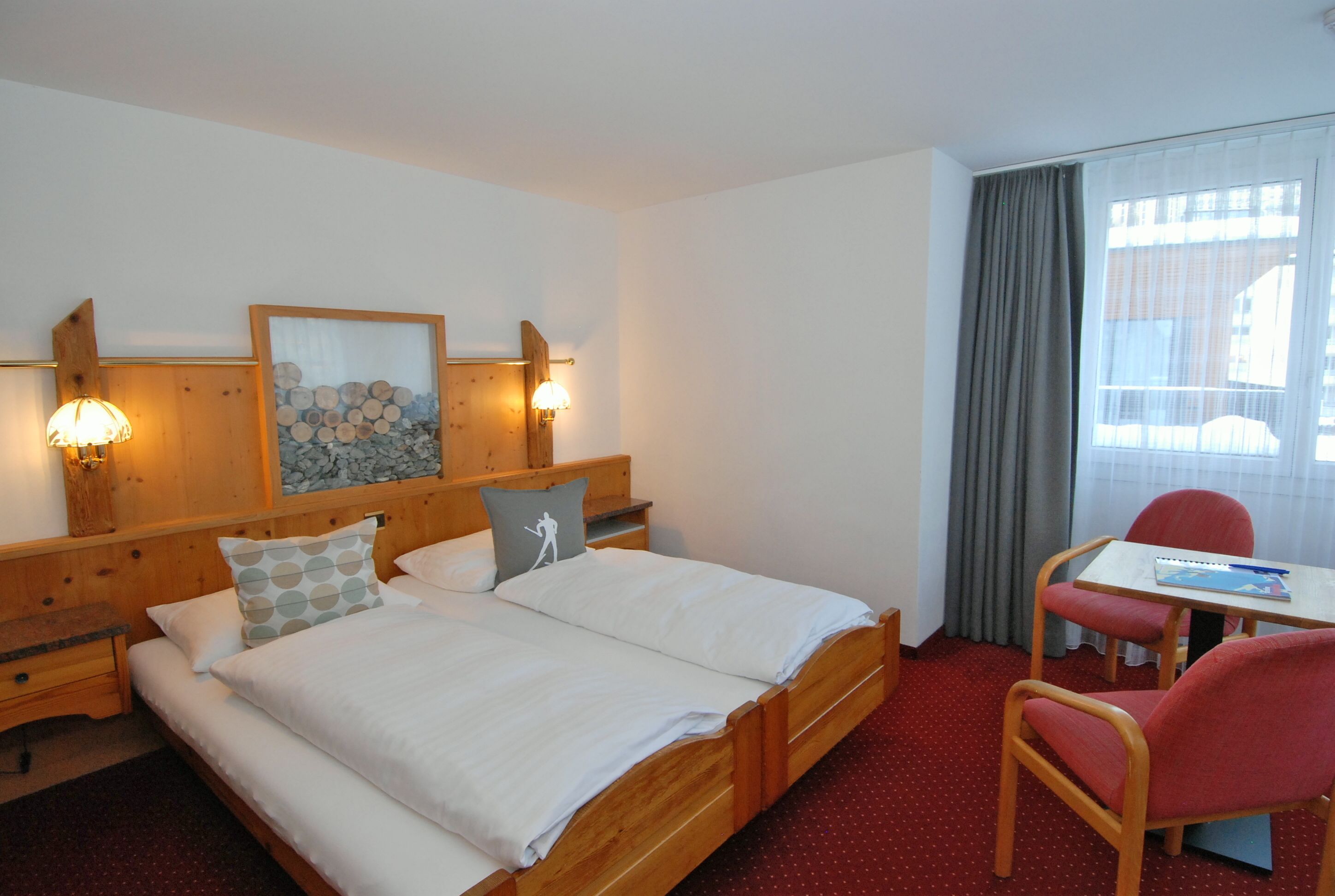 A hotel room with a wooden bed, two bedside tables, a desk with a chair, and a window with curtains.