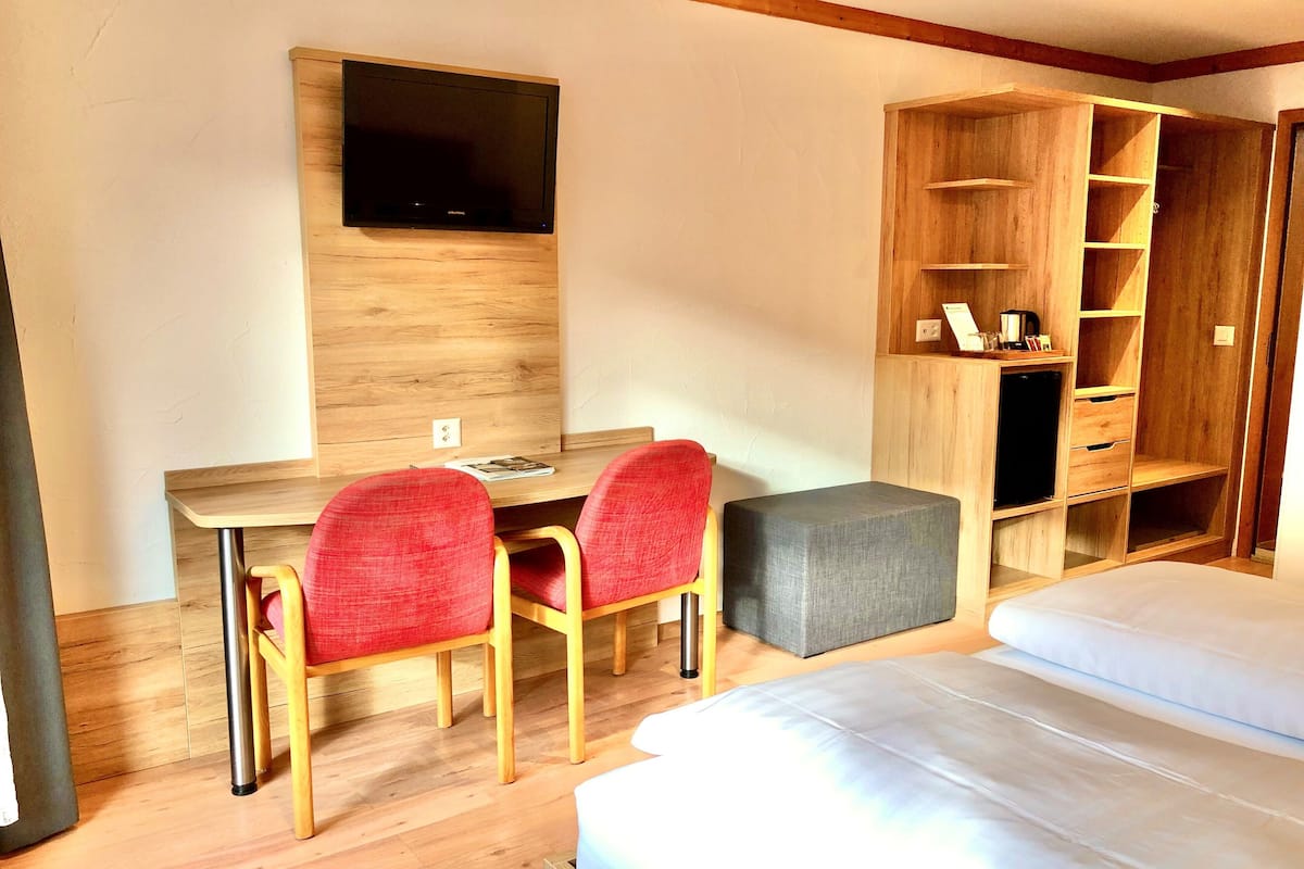 Une chambre d’hôtel avec un bureau en bois, deux chaises rouges, un téléviseur fixé au mur, une armoire avec plusieurs étagères et un lit avec une literie blanche.