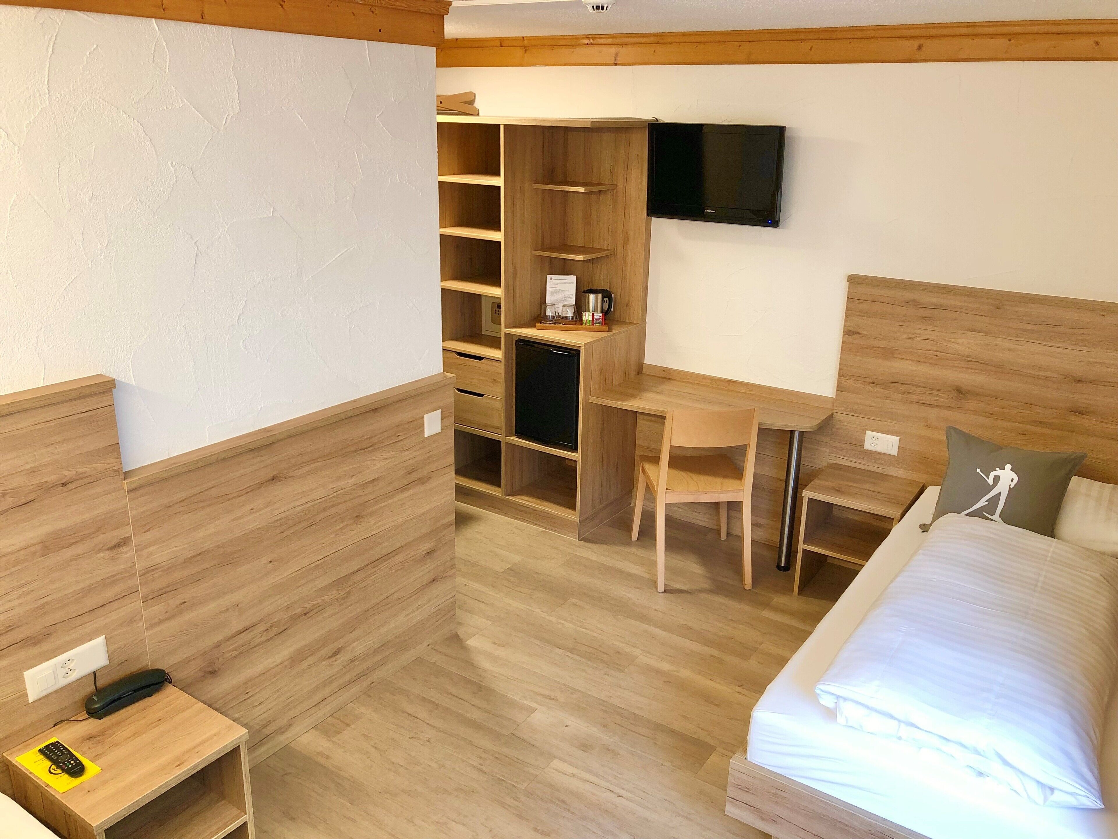 A hotel room with a wooden floor, a bed, a desk with a chair, a TV mounted on the wall, and a wardrobe with shelves.