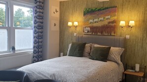 Een slaapkamer met een groot bed, twee wandlampen, een schilderij van schapen aan de muur en een raam met gordijnen.