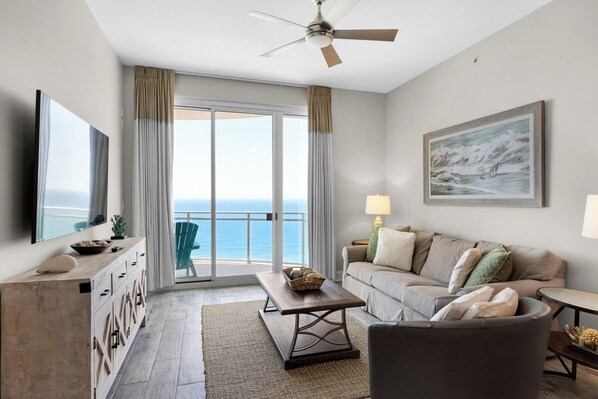 A modern living room with a sofa, coffee table, and TV, a balcony with a view of the ocean, and a ceiling fan.