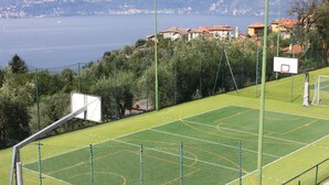 An outdoor basketball court with a net, surrounded by a green fence, overlooking a lake with buildings in the background. - Goldenlake Apartment (Perledo)