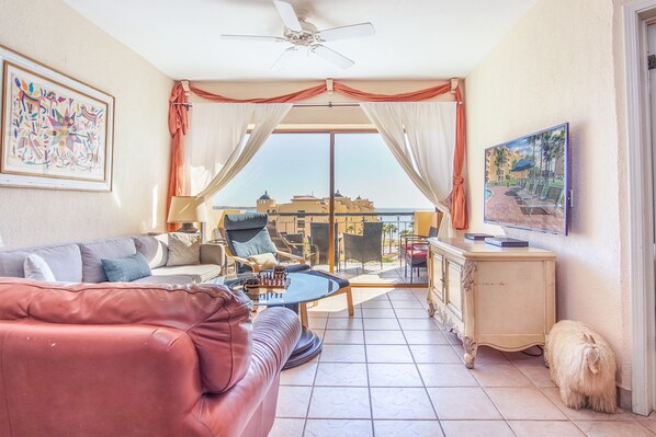 Un salon avec un canapé, une table basse et un téléviseur. Il y a un balcon d’où l’on peut admirer les bâtiments et la mer.