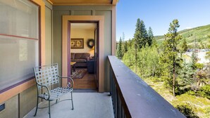 Un balcon avec un banc en métal, une porte donnant sur une pièce avec un canapé et un tapis, et une vue sur une colline boisée.