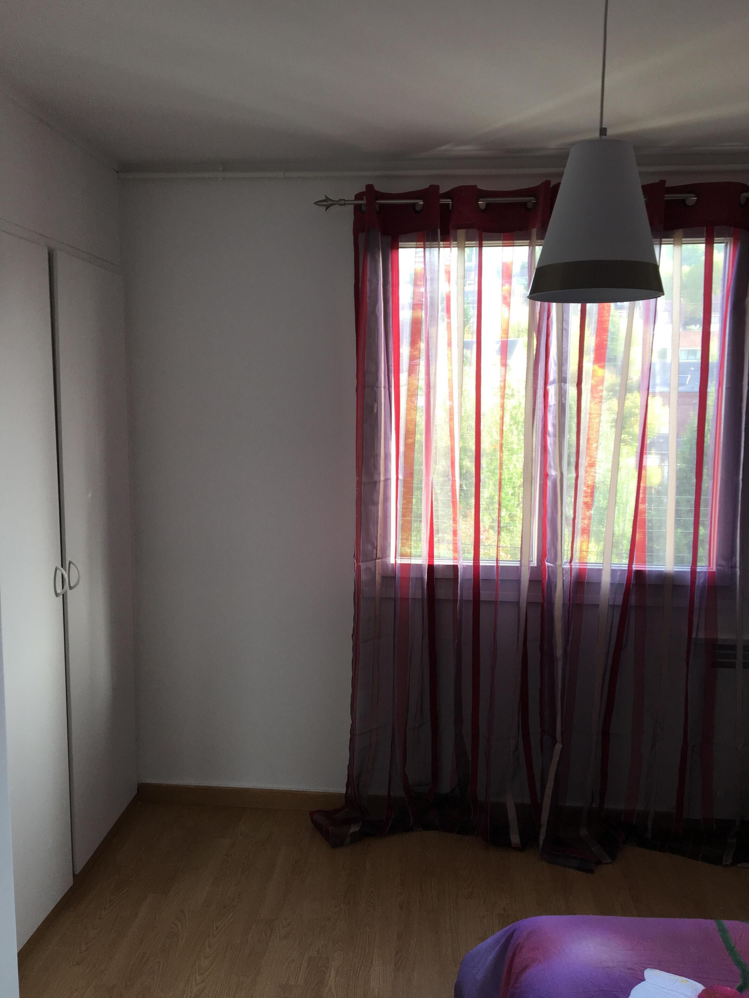 Une pièce avec un plancher en bois, un lit recouvert d’une courtepointe violette, une fenêtre ornée de rideaux rouges et blancs, et une lampe suspendue.