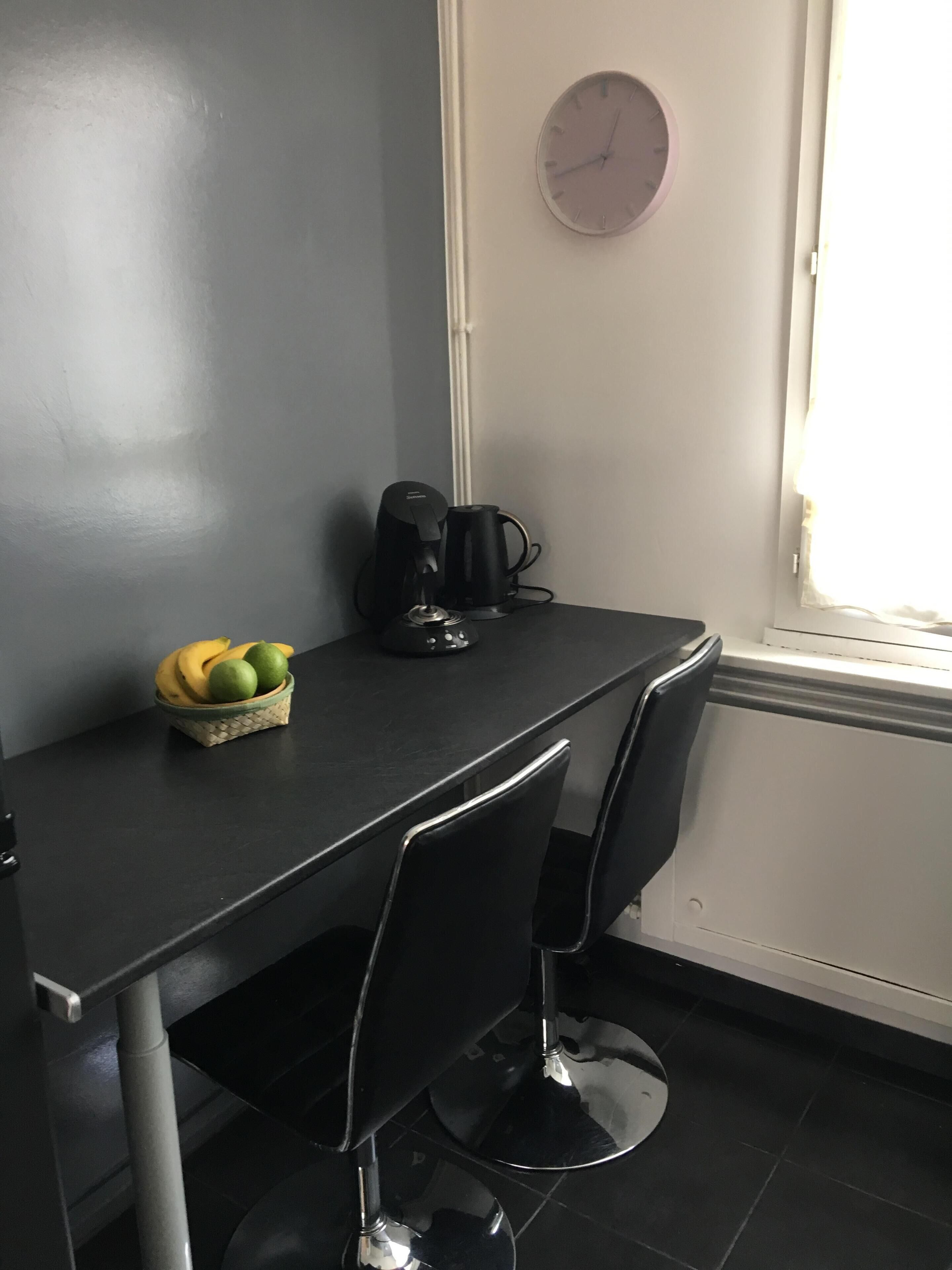A small, dark-colored table with a basket of fruit, a coffee maker, and a clock on the wall.