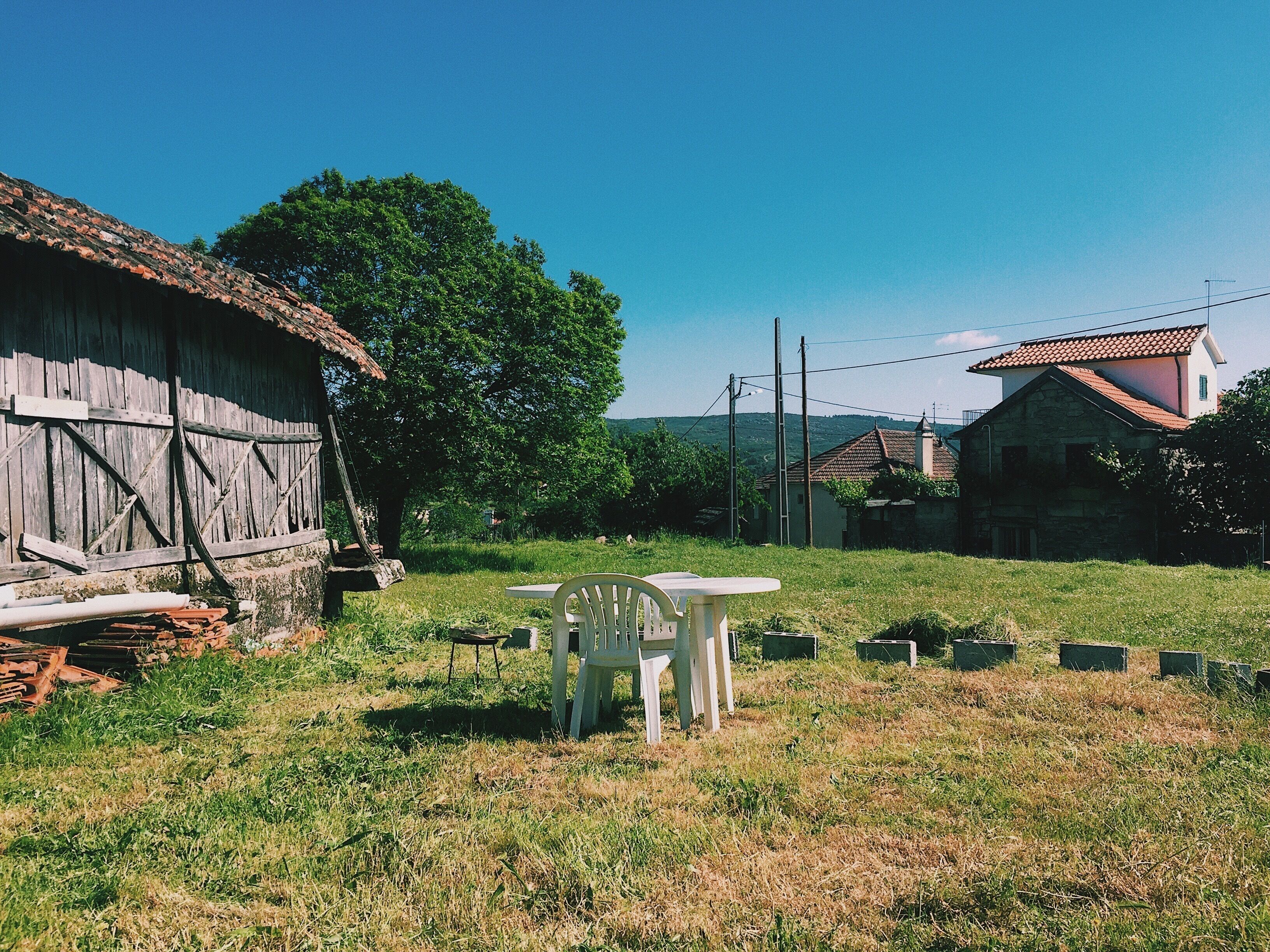 Entorno rural con una casa de madera, una mesa y una silla, y un cielo azul despejado.
