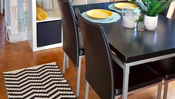 A dining area with a TV on a white stand, a black table, and chairs. There is a patterned rug on the floor.