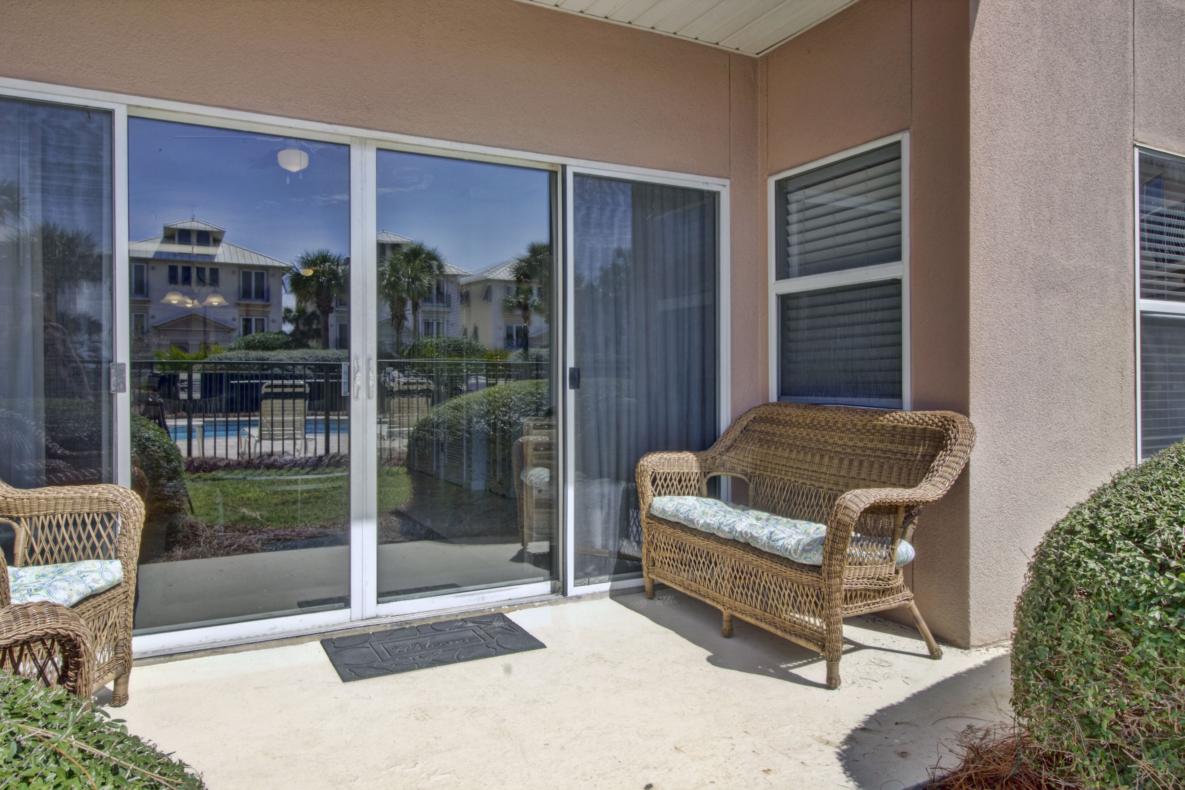 Un patio cubierto con mobiliario de mimbre, un gran ventanal corredizo y vistas a una piscina y casas al fondo.