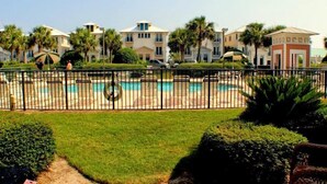 A pool area with a fence, surrounded by manicured lawns and palm trees, with buildings in the background. - Great Location- Heart of Miramar Beach! So Close to Pool, Beach, & More! (Miramar Beach)