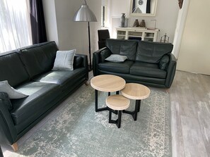 Ett modernt vardagsrum med en svart lädersoffa, två runda träbord och en stor golvlampa.