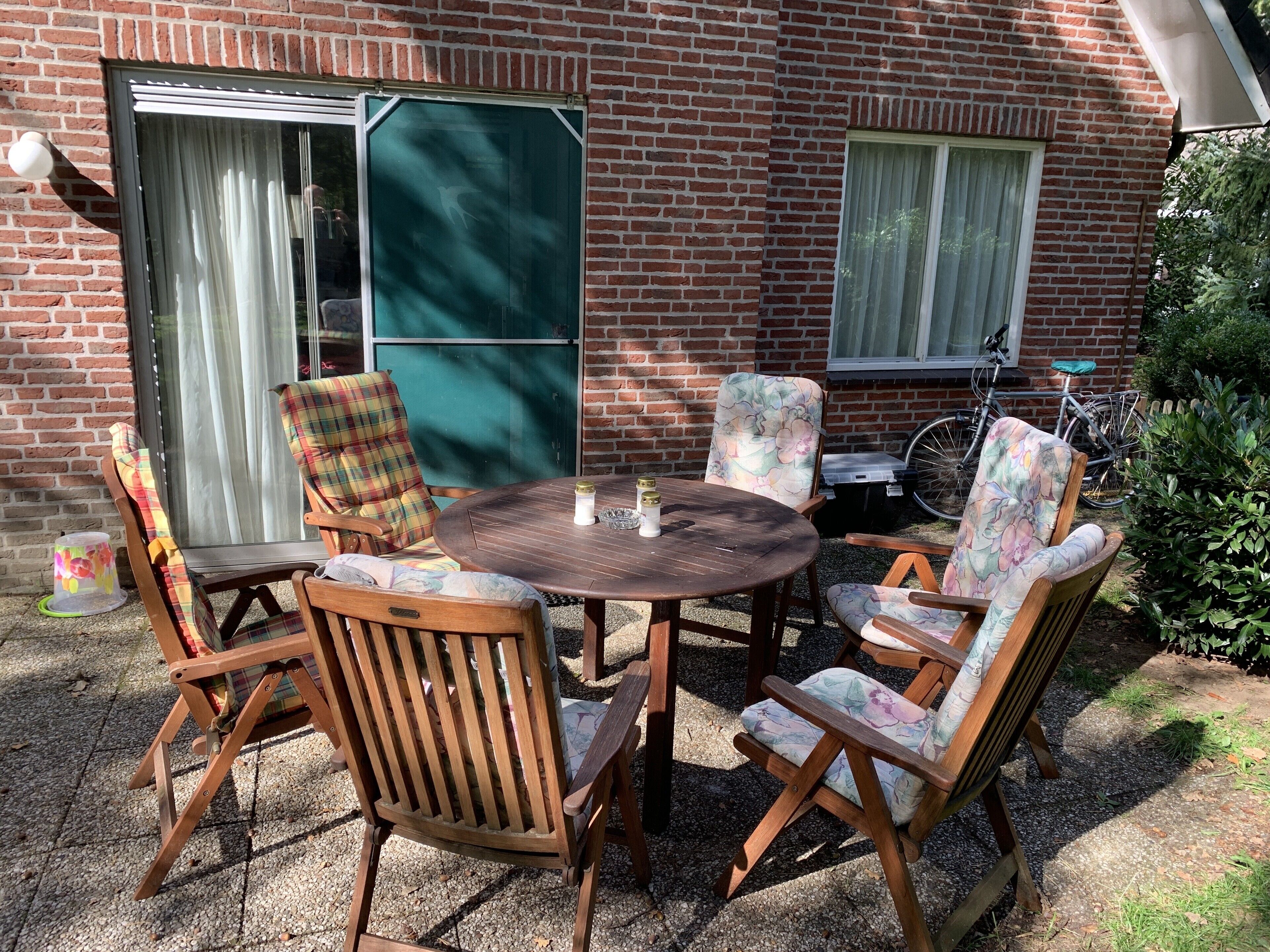 Une terrasse avec une table ronde en bois et quatre chaises, un vélo, et un bâtiment en briques doté d’une porte-fenêtre coulissante.