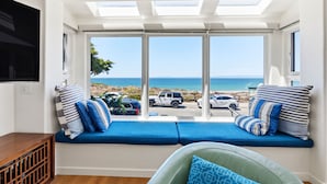 Una habitación con un amplio ventanal que da a una playa, un banco empotrado con cojines y un televisor fijado a la pared.