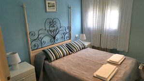 Una camera da letto con un letto, due comodini, una lampada, una finestra con tende in pizzo e un quadro appeso al muro.