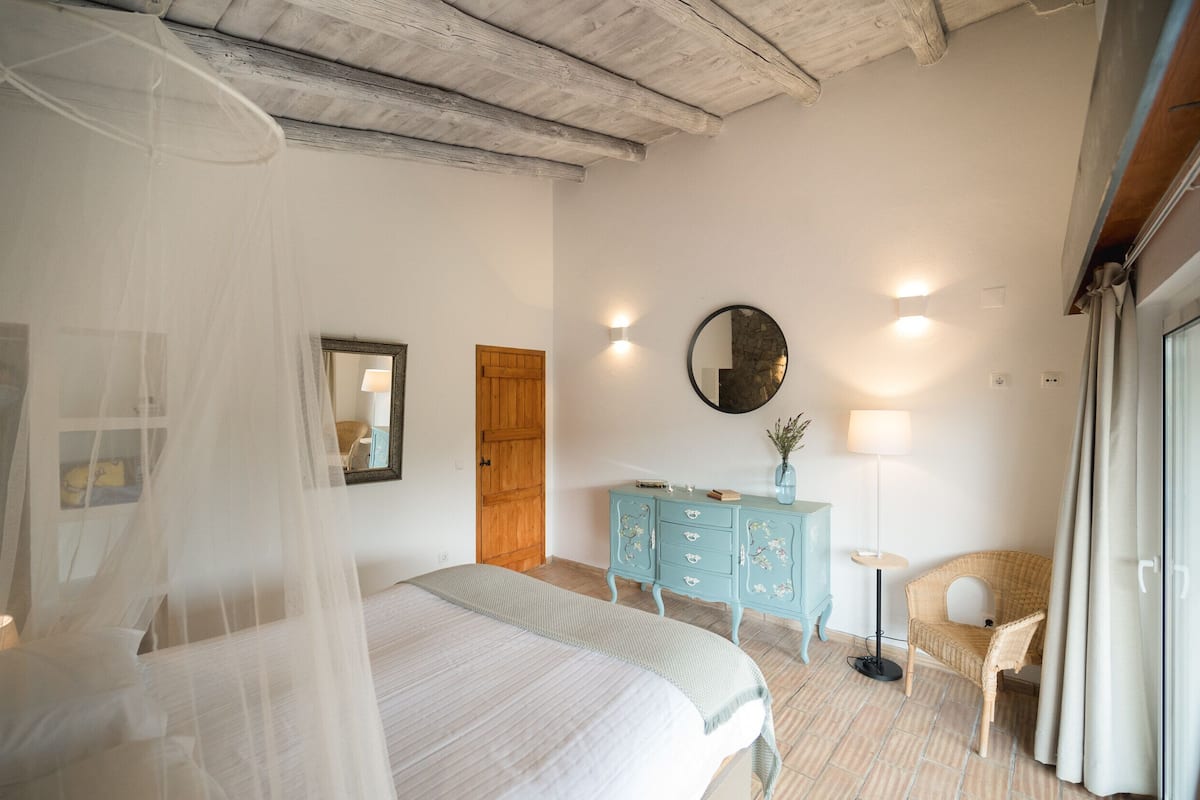 A bedroom with a bed, a wooden door, a blue dresser, a chair, a round mirror, and a nightstand with a lamp.