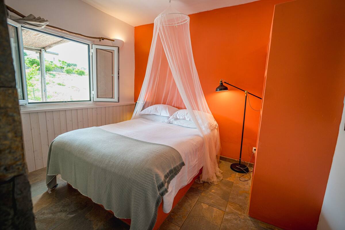 A bedroom with a bed under a canopy, a bedside lamp, and a window with a view of greenery.
