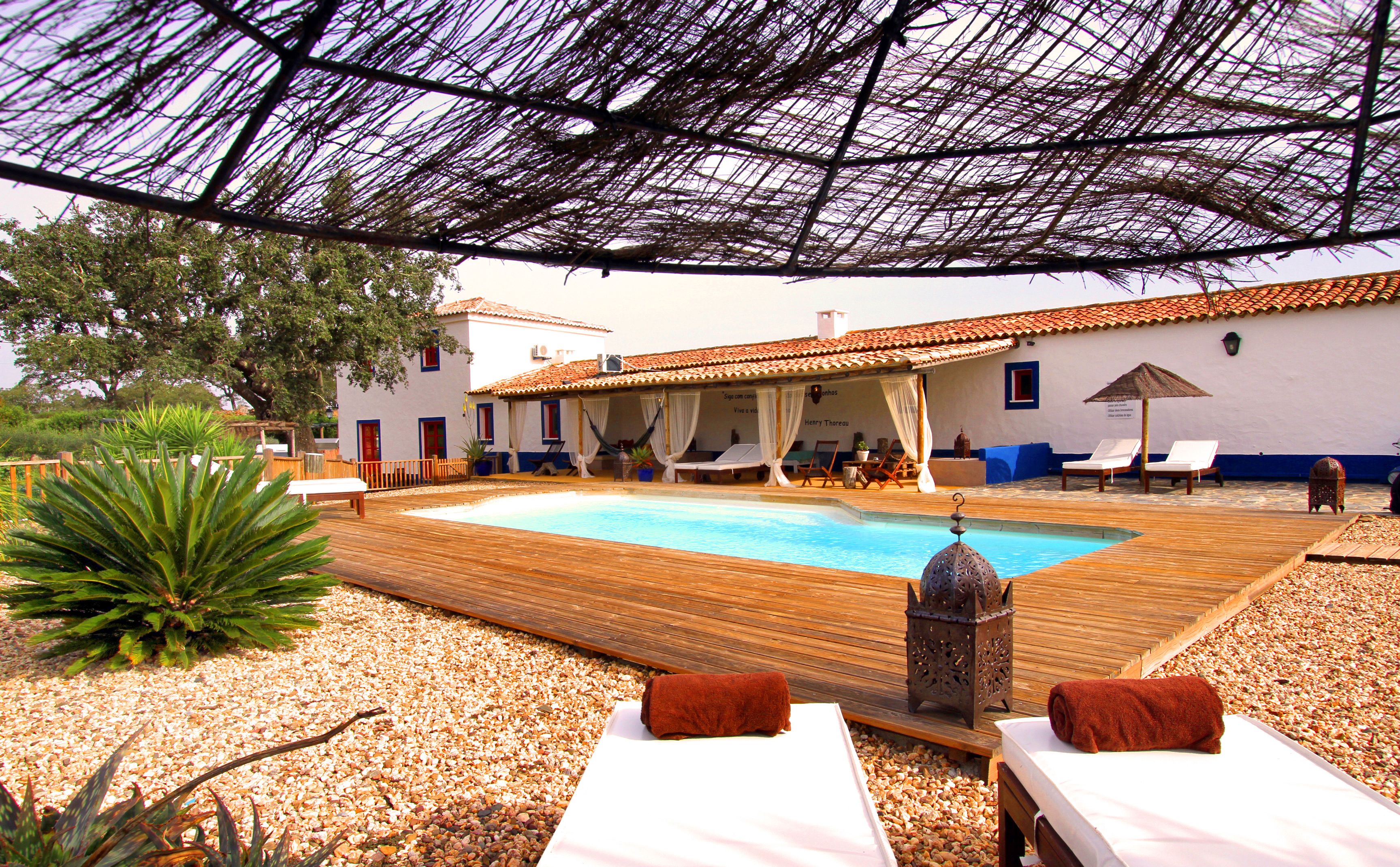 Un área de piscina con sillones de descanso, una terraza de madera y un edificio con tejado de azulejos.