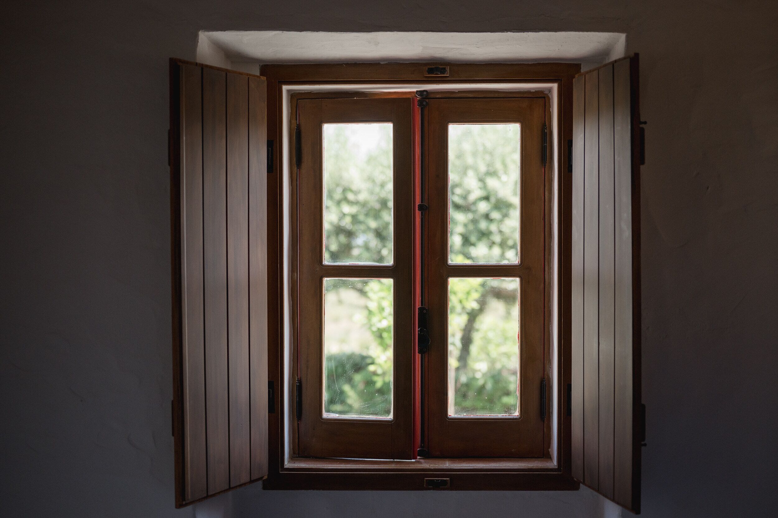 Una habitación con ventana de madera, con cuatro divisiones, y vista a la vegetación.