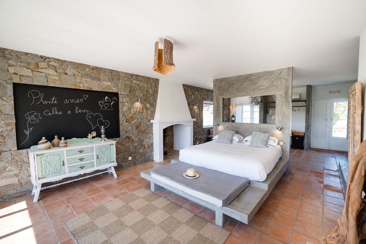 A bedroom with a stone wall, a white bed, a dresser, and a fireplace.
