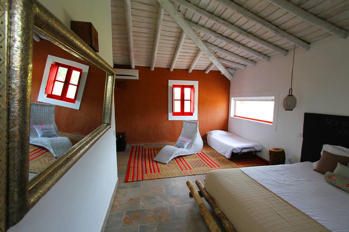 A bedroom with a bed, a chair, a small table, a mirror, a window, and a ceiling with wooden beams.