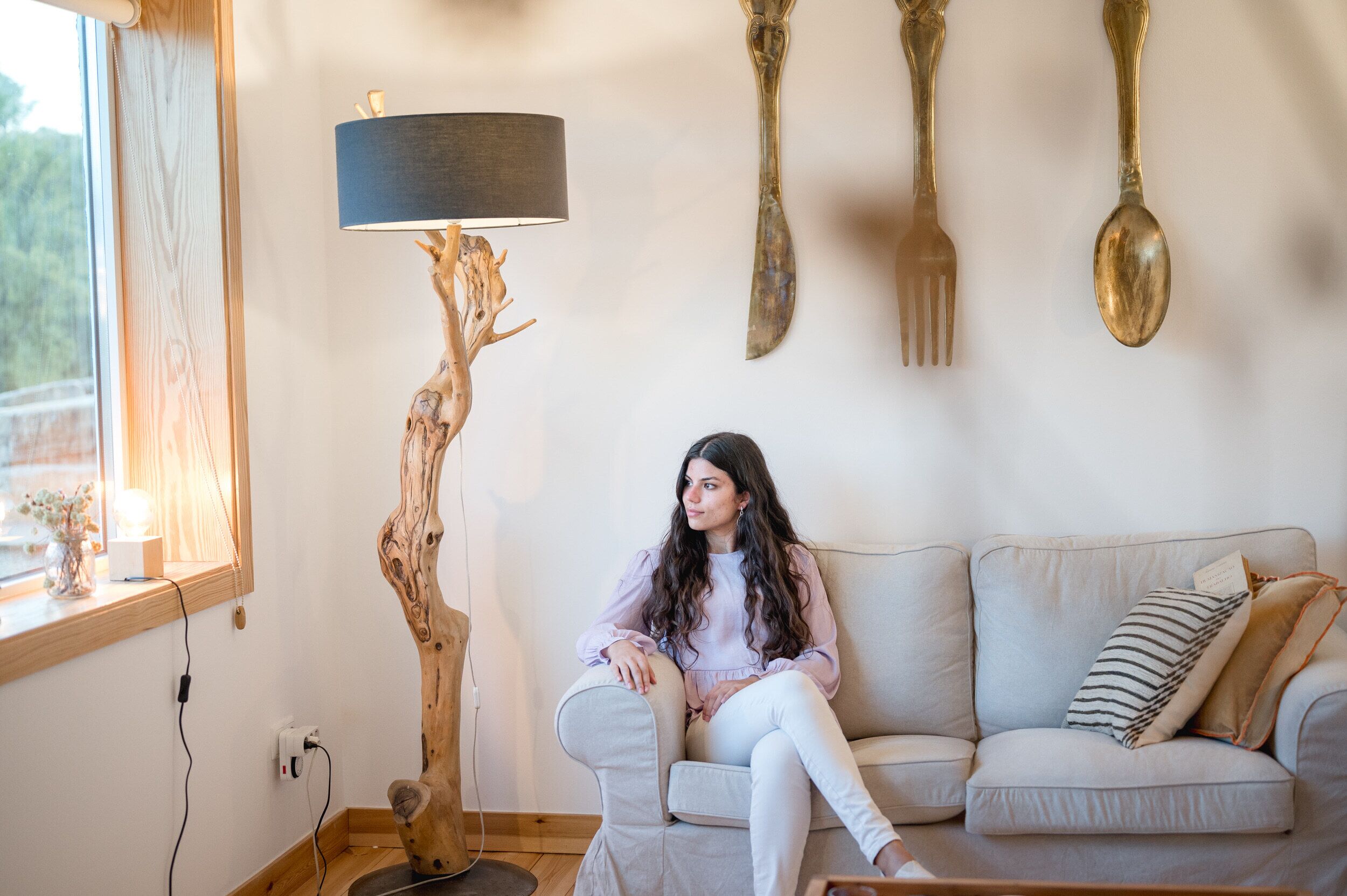 Una persona seduta su un divano in una stanza con una lampada da terra in legno, una finestra e posateria a parete.