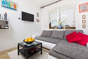 Una sala de estar con un sofá gris, una mesa de centro negra con un cuenco de fruta, un tapete blanco y una ventana con cortinas traslúcidas.