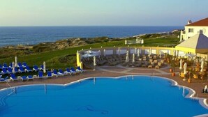Área de piscina com cadeiras de descanso azuis e guarda-sóis, um edifício com fachada amarela e vista para o mar.