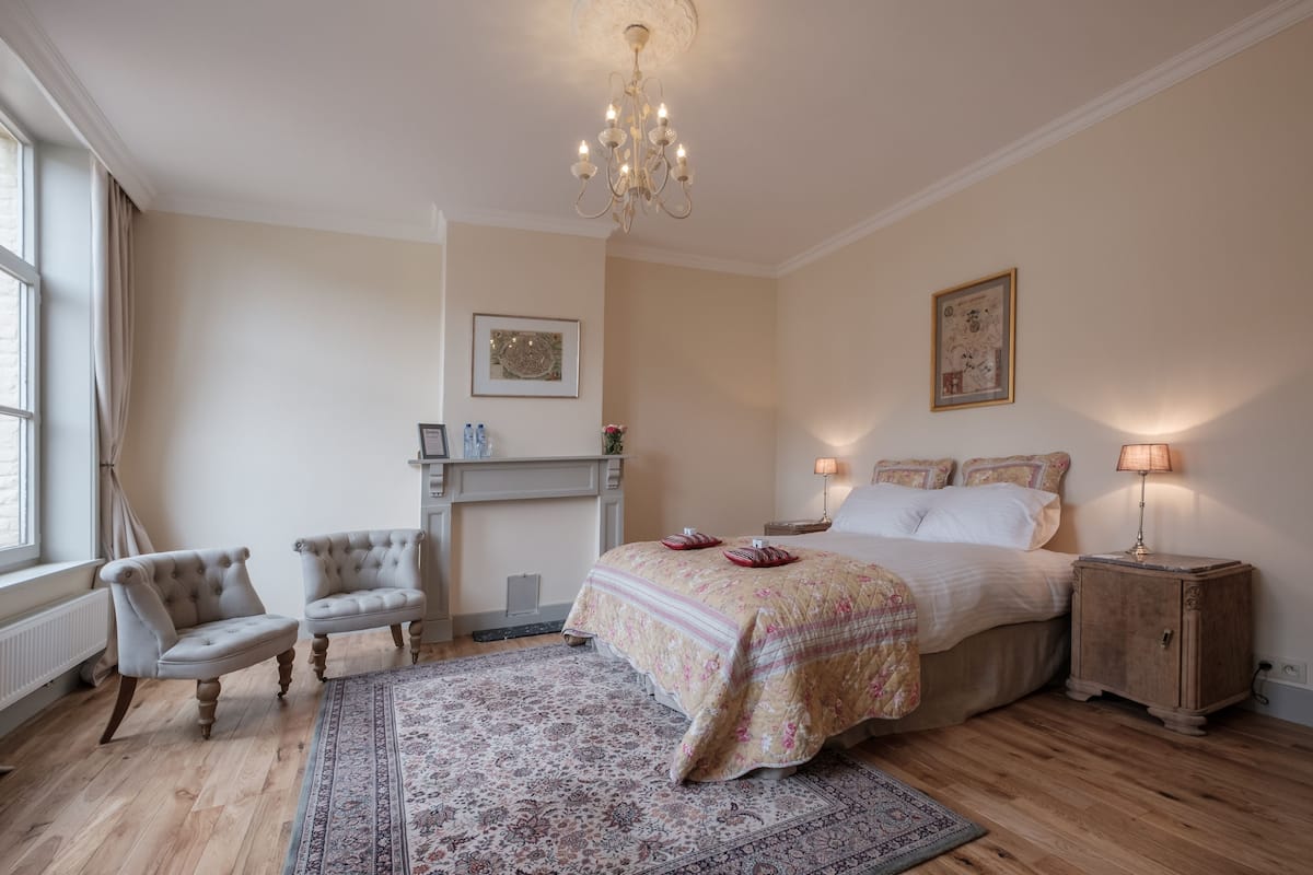 A bedroom with a large bed, two armchairs, a wooden floor, a chandelier, and a fireplace.
